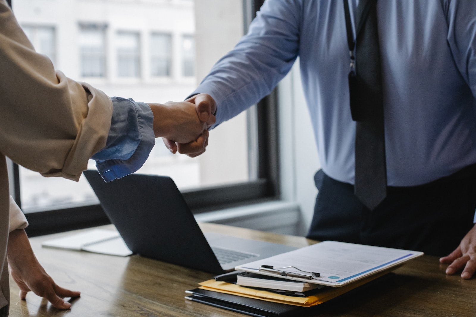 Photo by Sora Shimazaki Crop unrecognizable coworkers in formal wear standing at table with laptop and documents while greeting each other before meeting